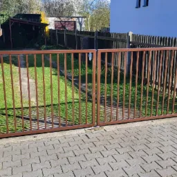 Nowoczesna, brązowa brama wjazdowa z metalowymi prętami, otwierana na posesję z kostką brukową, trawnikiem i zabudowaniami w tle.