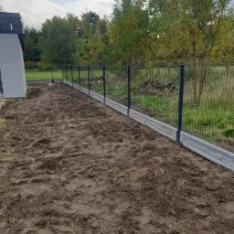 Montaż ogrodzenia panelowego z podmur&oacute;wką betonową na wilgotnej, przygotowanej ziemi, widok wzdłuż linii ogrodzenia, w tle budynek i drzewa.