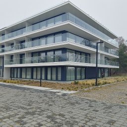 Chmielewski-Nieruchomości - Nowoczesny apartamentowiec z balkonami i szklanymi balustradami, otoczony brukowanym terenem i suchymi trawami, widok z poziomu gruntu w pochmurny dzień.