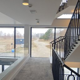 Chmielewski-Nieruchomości - Wnętrze nowoczesnego budynku z klatką schodową z czarnymi metalowymi balustradami, widok z antresoli na okna z widokiem na krajobraz z budynkami w tle.