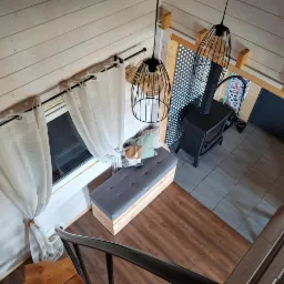 Wnętrze drewnianego domu z antresolą, widok z g&oacute;ry na piec, okno z zasłonami, ławkę z poduszkami i metalowe schody. Nowoczesny design, jasne kolory.