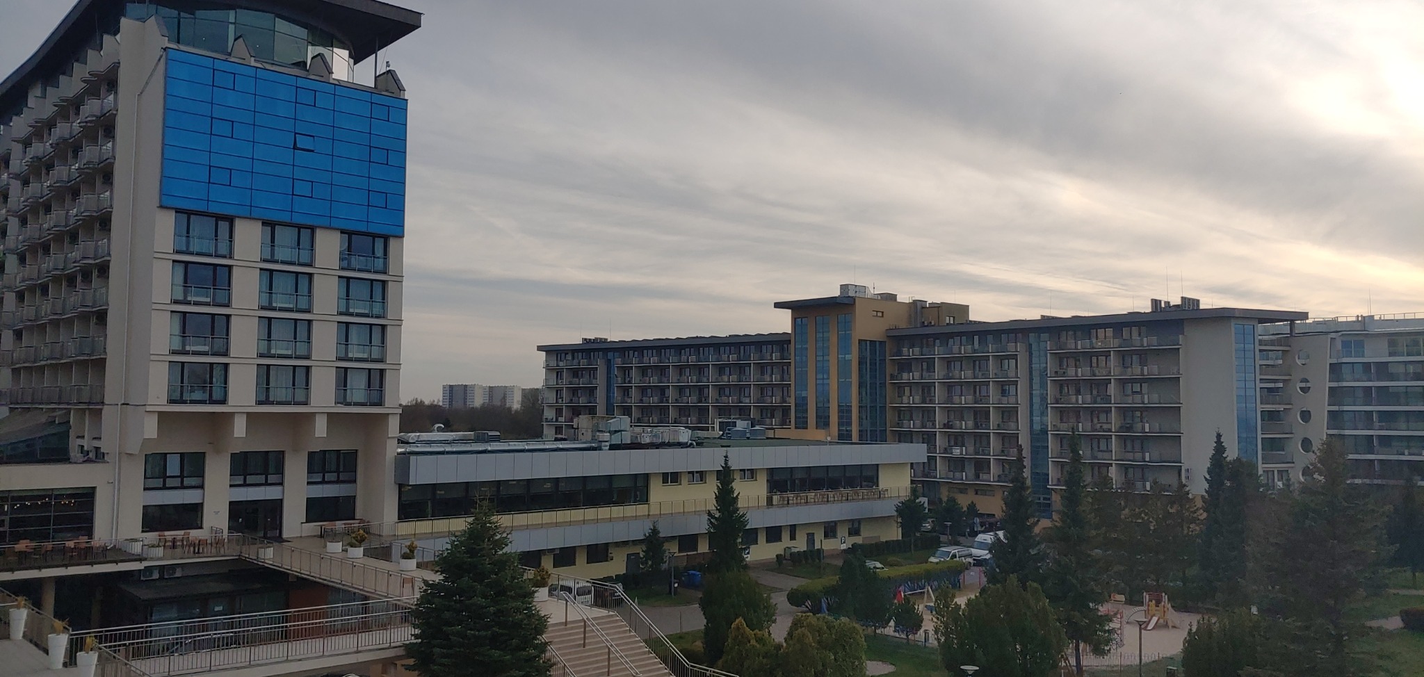 Widok z góry na kompleks apartamentowo-hotelowy z wyraźnym podziałem na sekcje o różnej architekturze i kolorystyce, otoczony zielenią drzew i krzewów, z parkingiem widocznym w tle, pod pochmurnym...