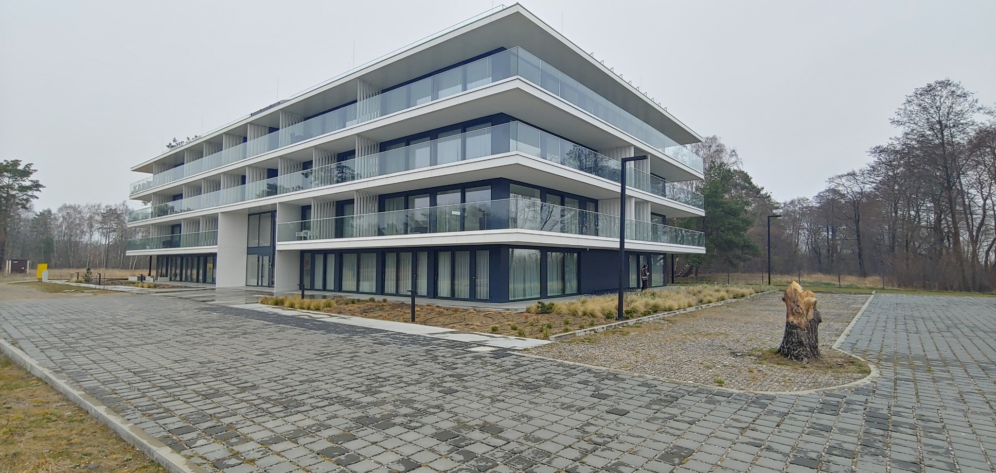 Nowoczesny apartamentowiec z balkonami i szklanymi balustradami, otoczony brukowanym terenem i suchymi trawami, widok z poziomu gruntu w pochmurny dzień.