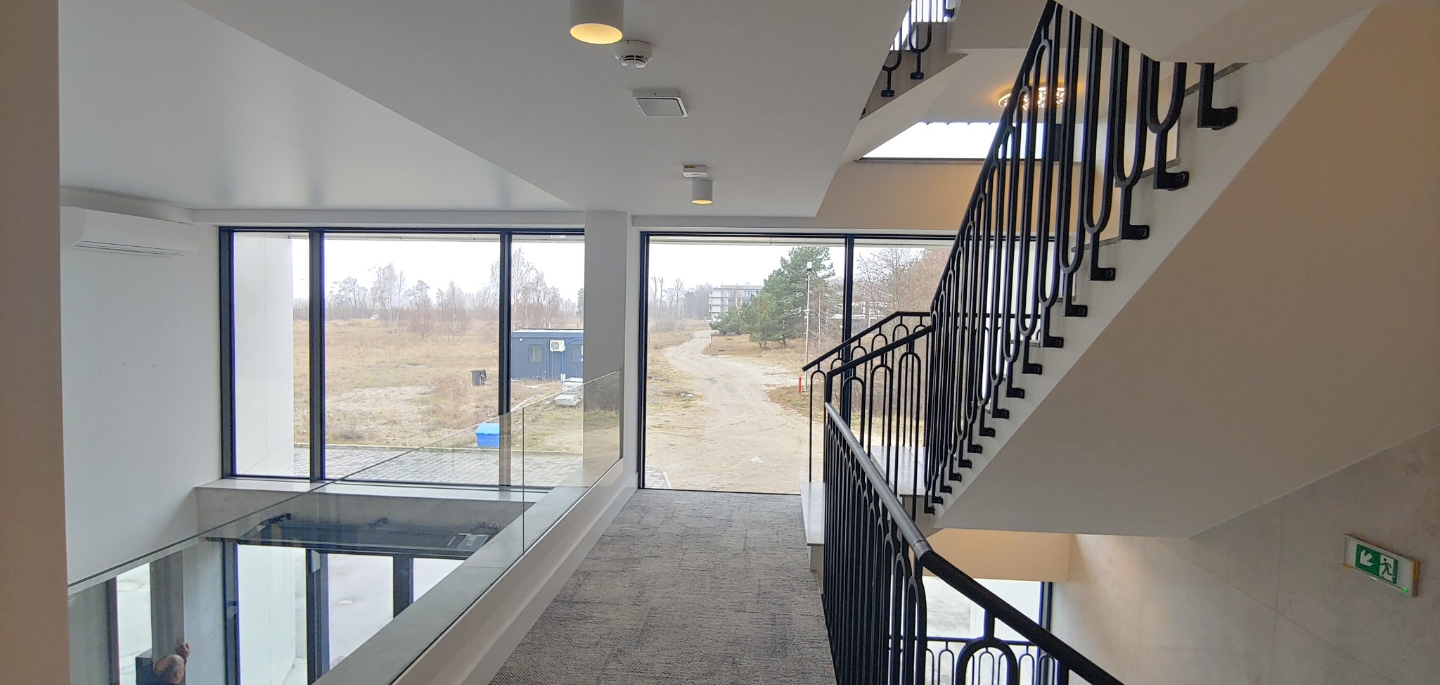 Wnętrze nowoczesnego budynku z klatką schodową z czarnymi metalowymi balustradami, widok z antresoli na okna z widokiem na krajobraz z budynkami w tle.
