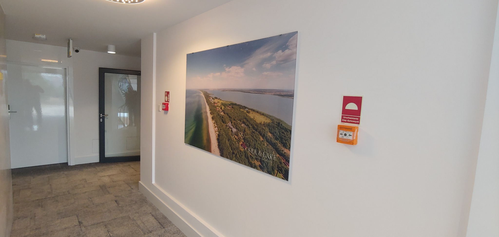 Korytarz w apartamentowcu z obrazem przedstawiającym widok na mierzeję i jezioro, z widocznymi drzwiami wejściowymi i systemem przeciwpożarowym.