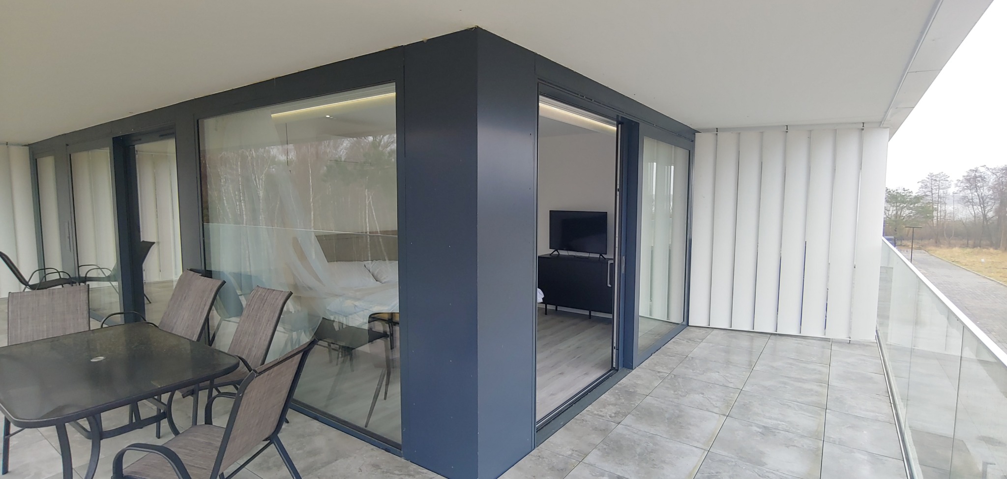 Przestronny balkon z szarymi ramami okiennymi i meblami ogrodowymi, widok na wnętrze apartamentu z telewizorem i łóżkiem