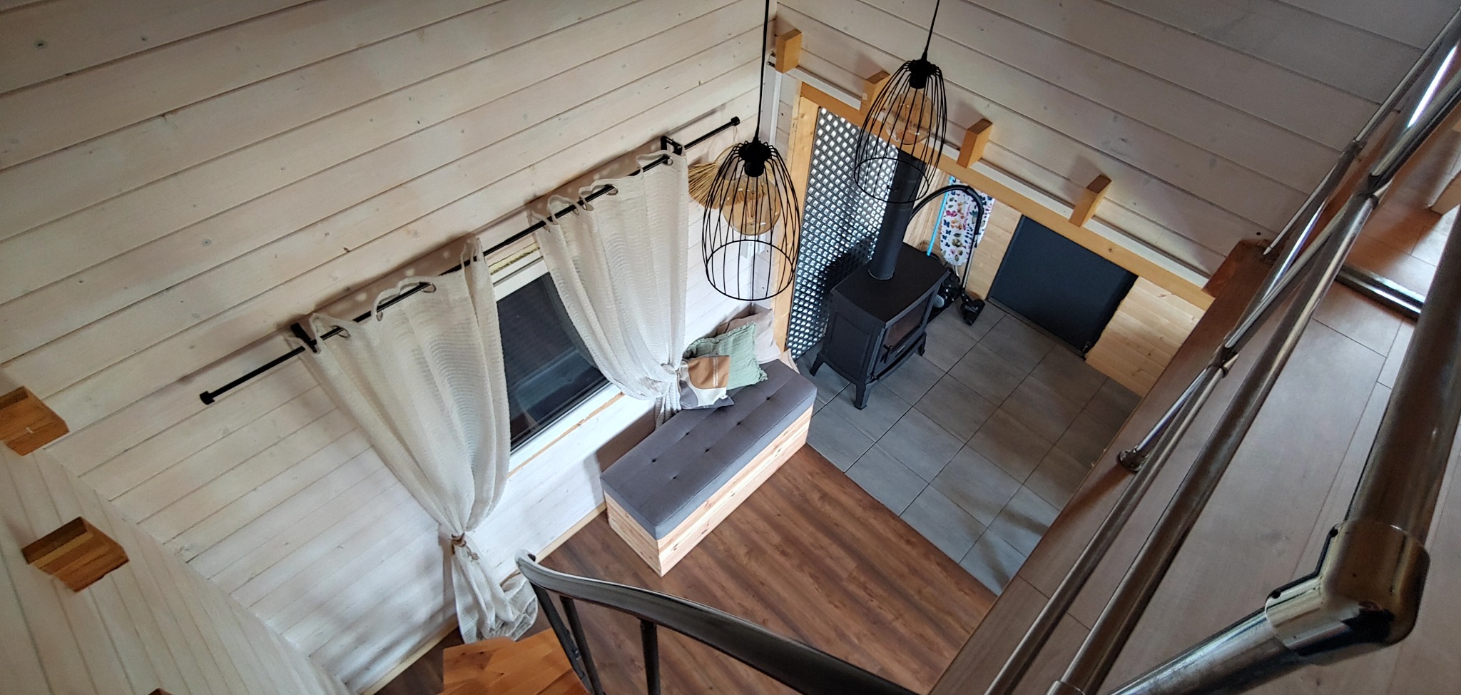 Wnętrze drewnianego domu z antresolą, widok z góry na piec, okno z zasłonami, ławkę z poduszkami i metalowe schody. Nowoczesny design, jasne kolory.