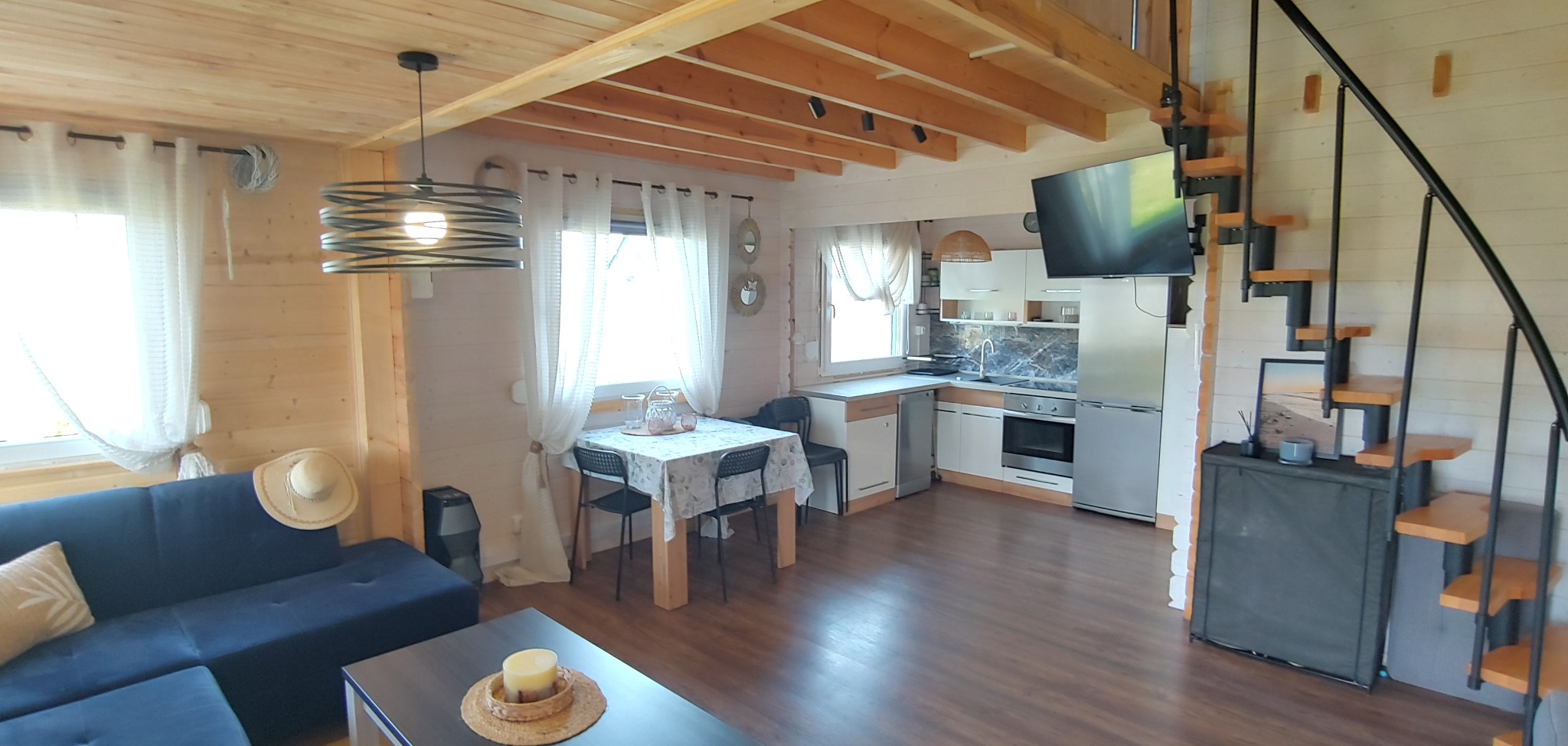 Jasne wnętrze domku letniskowego z drewnianymi elementami, spiralnymi schodami i aneksem kuchennym. Przytulny salon z kanapą i stolikiem kawowym.