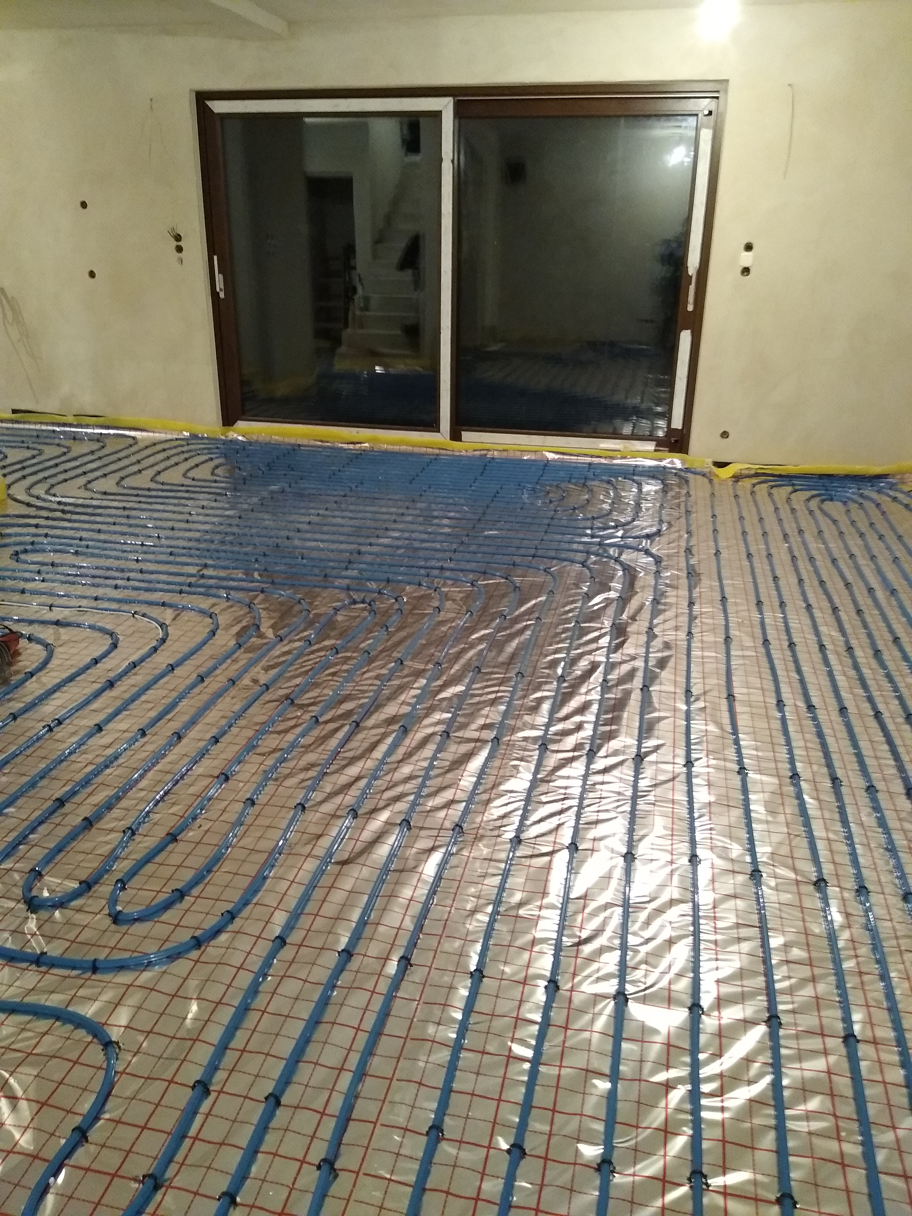 Niebieskie rury ogrzewania podłogowego ułożone na folii odblaskowej z czerwoną siatką, widok na surowe ściany i okno przesuwne w tle.