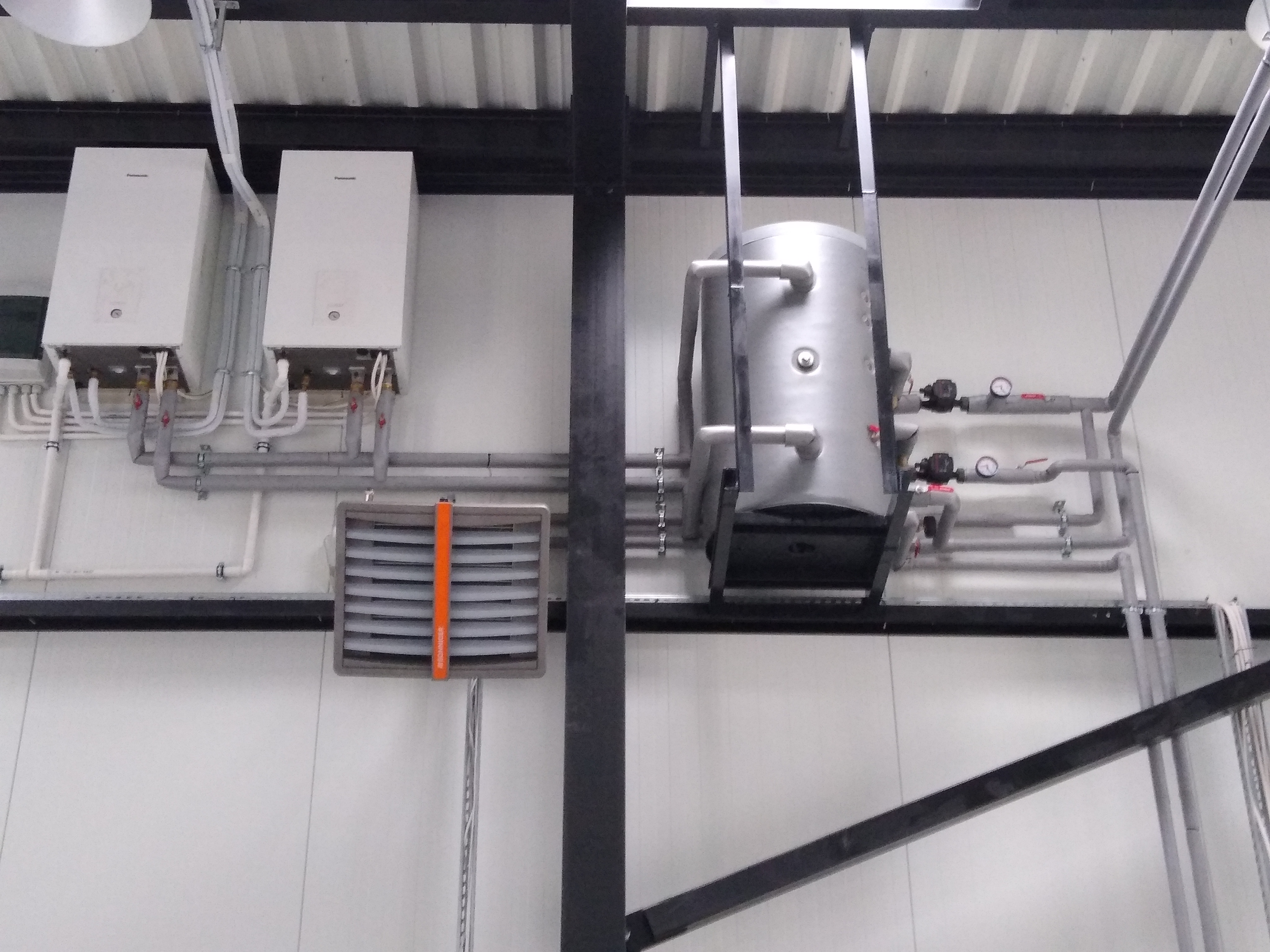 Dwa białe kotły grzewcze wiszące na ścianie, połączone rurami z szarym zbiornikiem akumulacyjnym, całość zamontowana w hali z blachy trapezowej.