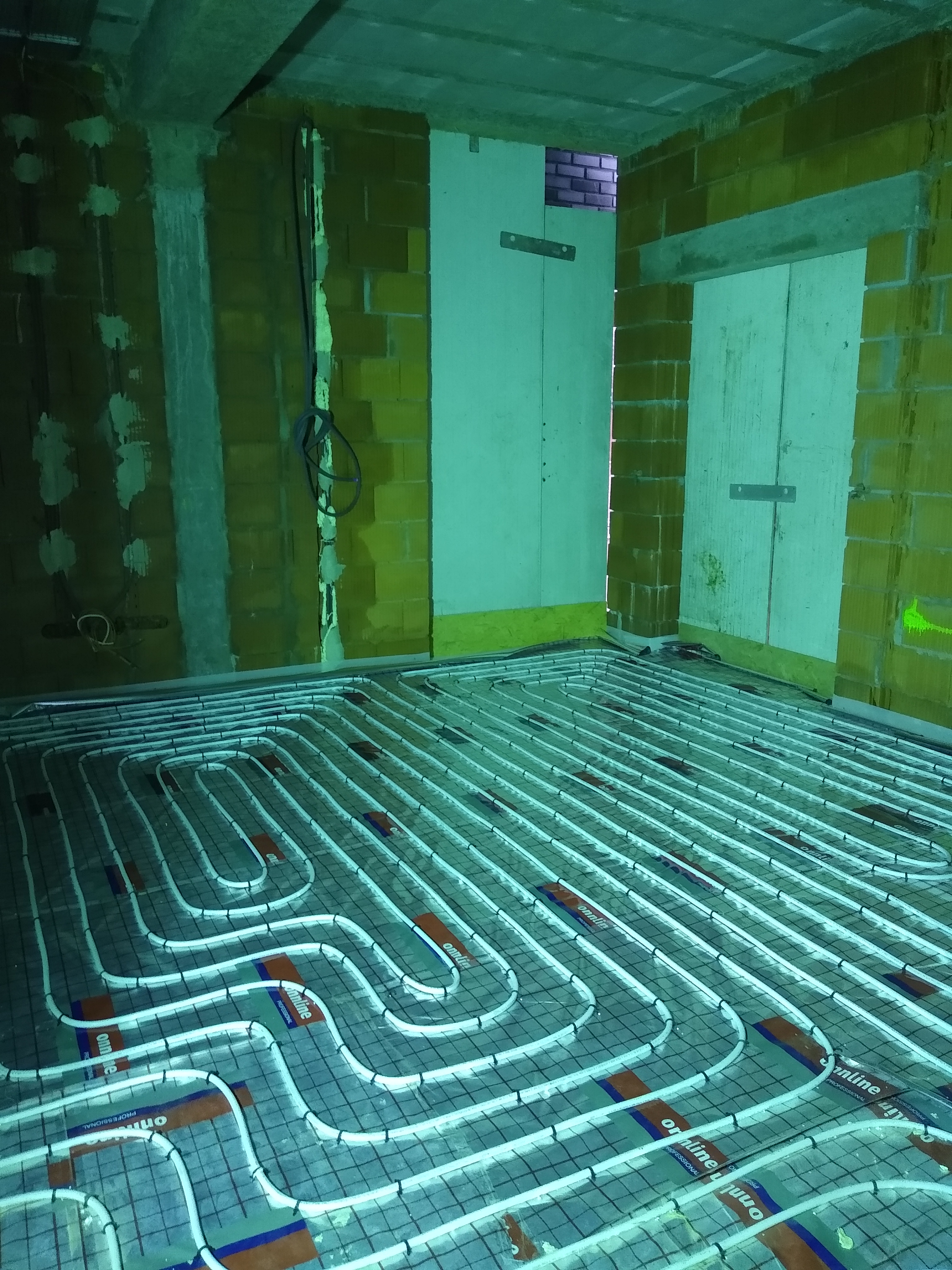 Instalacja podłogowa ogrzewania w surowym wnętrzu z ceglanymi ścianami, widoczne białe rury grzewcze ułożone na siatce montażowej, przygotowane do zalania wylewką.