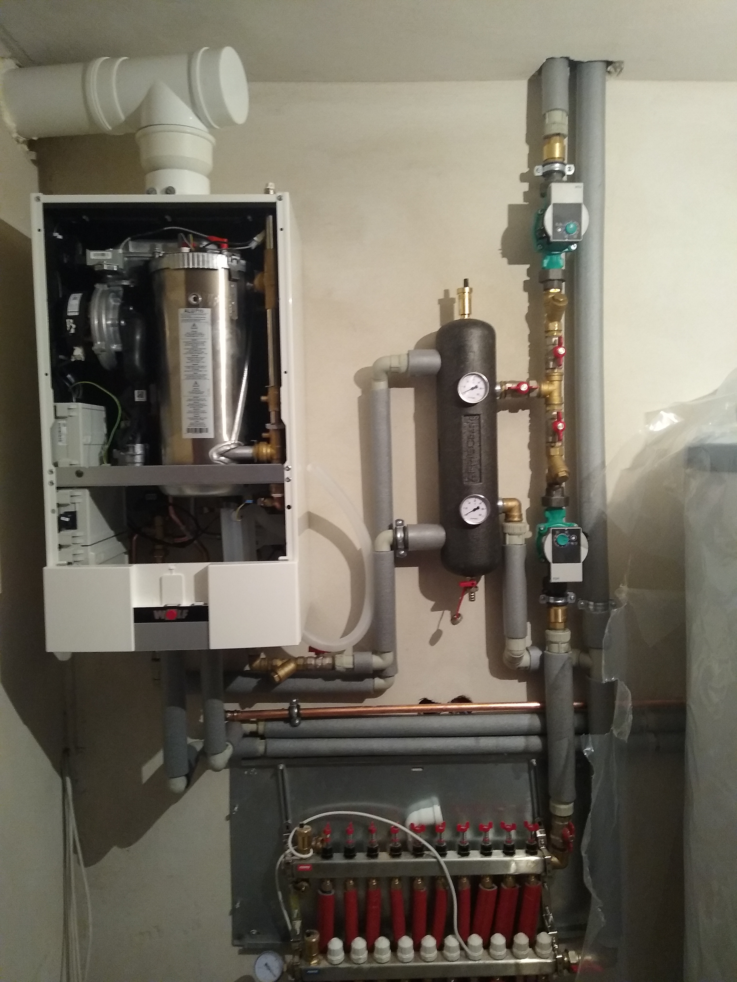 Zainstalowany system grzewczy z kotłem gazowym, zasobnikiem ciepła i rozdzielaczem podłogowym, widoczne rury i zawory, całość zamontowana na ścianie.