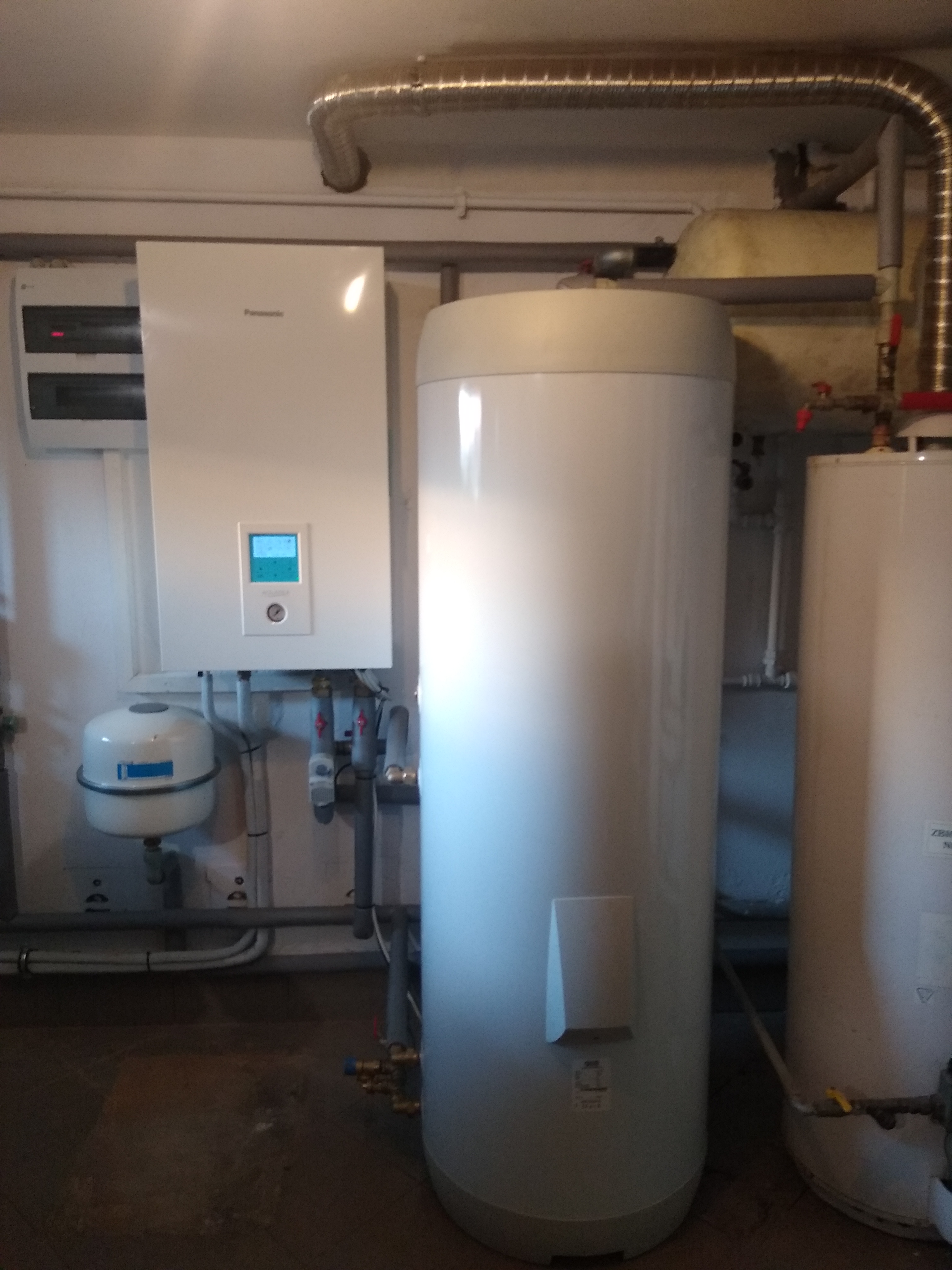 Biała jednostka wewnętrzna z wyświetlaczem i zbiornik buforowy w kotłowni, widoczne rury i instalacje.