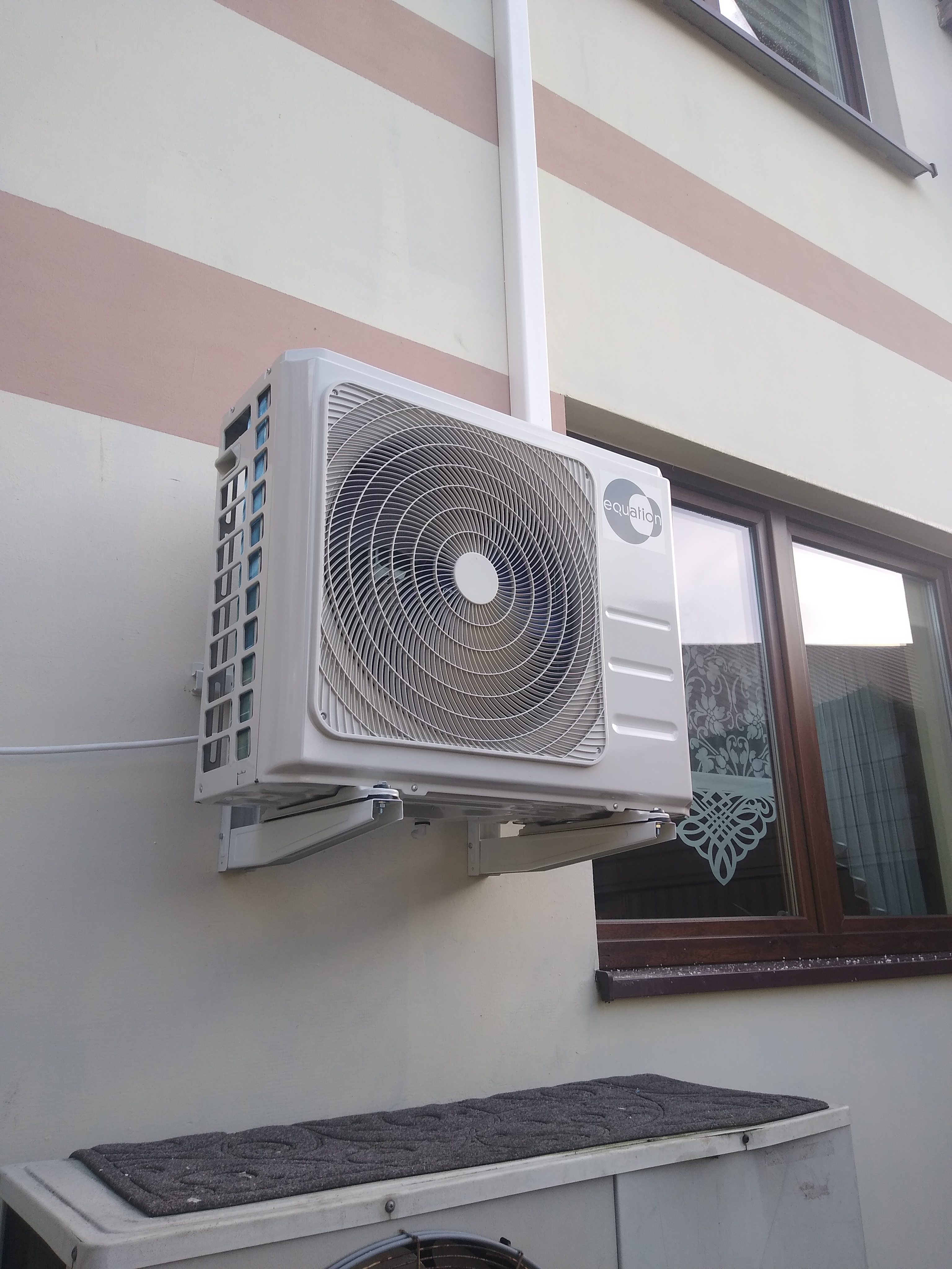 Biała jednostka zewnętrzna klimatyzacji marki Equation zamontowana na kremowej ścianie budynku z brązowymi pasami, widoczna rura odprowadzająca skropliny oraz okno z firanką.