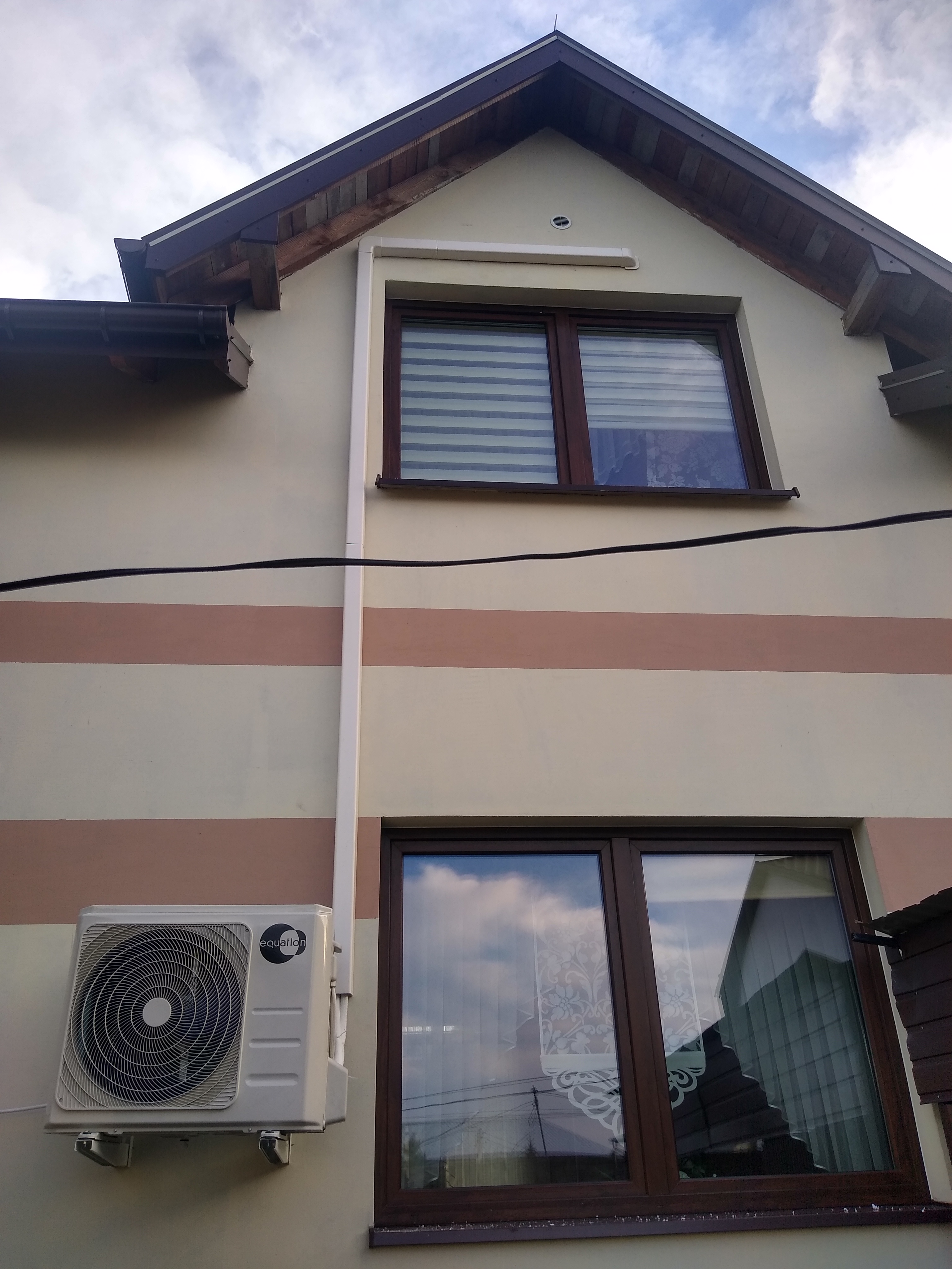 Zewnętrzna jednostka pompy ciepła Equation zamontowana na elewacji domu z oknami i brązowymi pasami, widoczna instalacja rur.