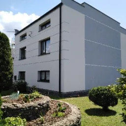 Dwukolorowa elewacja trzypiętrowego budynku mieszkalnego z ogrodem skalnym w pierwszym planie, widok z zewnątrz w słoneczny dzień.