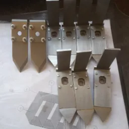 Sześć metalowych elementów z ostrą końcówką, przymocowanych śrubami do płaskich płytek, ułożonych na tle schematu technicznego.