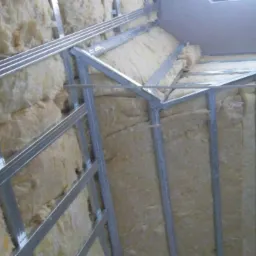 Skośne poddasze w trakcie ocieplania wełną mineralną, widoczne metalowe profile konstrukcyjne, surowe wykończenie, widok z dołu, oświetlenie g&oacute;rne.