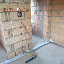 Wnętrze w trakcie prac elektrycznych: kable w ścianach z cegły, puszki instalacyjne, przewody pod sufitem, miotła na podłodze. Instalacja elektryczna w surowym stanie.