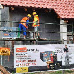 DeKaWu Grzegorz Olczyk - Pracownicy na rusztowaniu podczas nakładania warstwy izolacyjnej na ścianę domu z widocznym banerem reklamowym firmy Termo Organika oraz tablicą ostrzegawczą teren budowy.