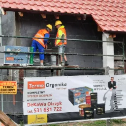 Pracownicy na rusztowaniu podczas nakładania warstwy izolacyjnej na ścianę domu z widocznym banerem reklamowym firmy Termo Organika oraz tablicą ostrzegawczą teren budowy.
