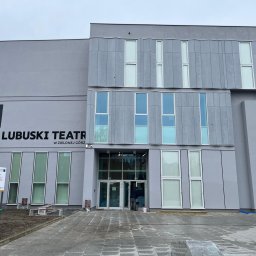 DeKaWu Grzegorz Olczyk - Szary budynek Lubuskiego Teatru w Zielonej Górze z nowoczesną elewacją, widoczne okna o różnych rozmiarach i kształtach, oraz częściowo zagospodarowany teren przed wejściem.
