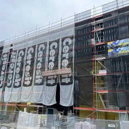 DeKaWu Grzegorz Olczyk - Ocieplanie elewacji budynku wielorodzinnego z użyciem rusztowań, osłonięte banerami reklamowymi Cerestit, widoczne płyty izolacyjne i siatka pod tynk.