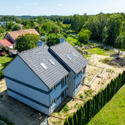 Dwukondygnacyjny dom murowany z szarą elewacją w trakcie budowy, widok z lotu ptaka, z fundamentami i otaczającą zielenią.