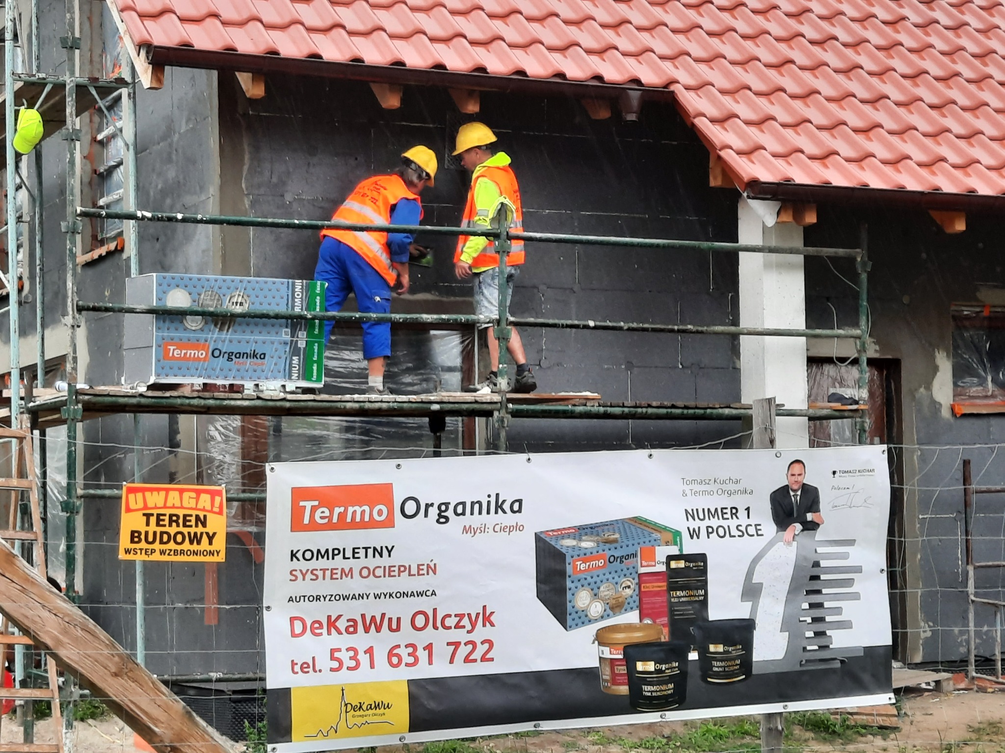 Pracownicy na rusztowaniu podczas nakładania warstwy izolacyjnej na ścianę domu z widocznym banerem reklamowym firmy Termo Organika oraz tablicą ostrzegawczą teren budowy.