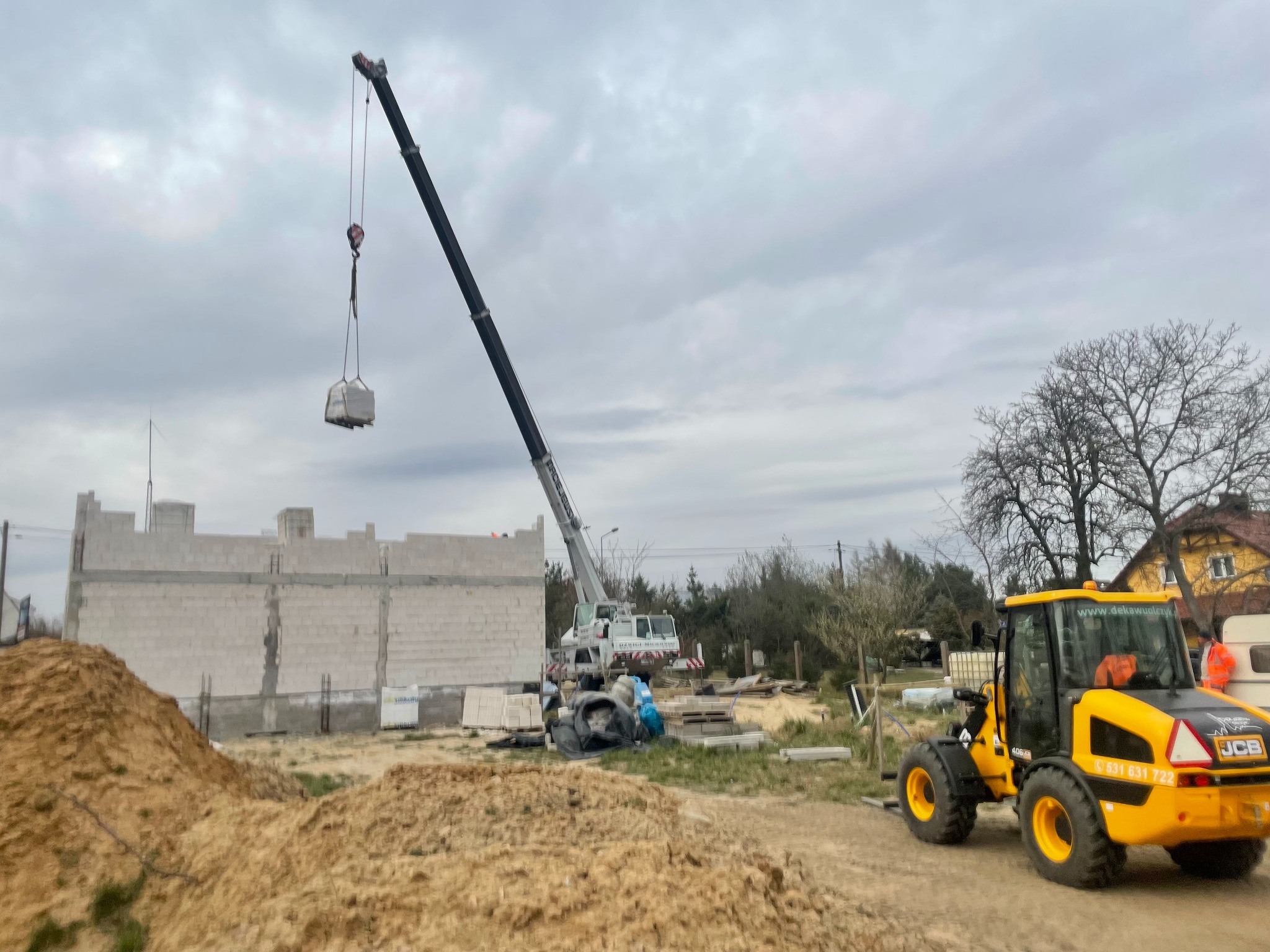 Budowa domu z białych bloczków: żuraw podnosi paletę z materiałem, na pierwszym planie żółta ładowarka JCB, w tle częściowo wzniesione ściany budynku.