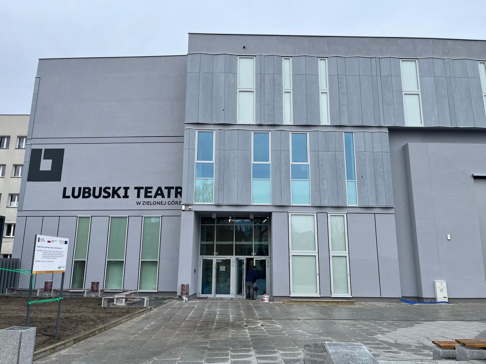 Szary budynek Lubuskiego Teatru w Zielonej Górze z nowoczesną elewacją, widoczne okna o różnych rozmiarach i kształtach, oraz częściowo zagospodarowany teren przed wejściem.