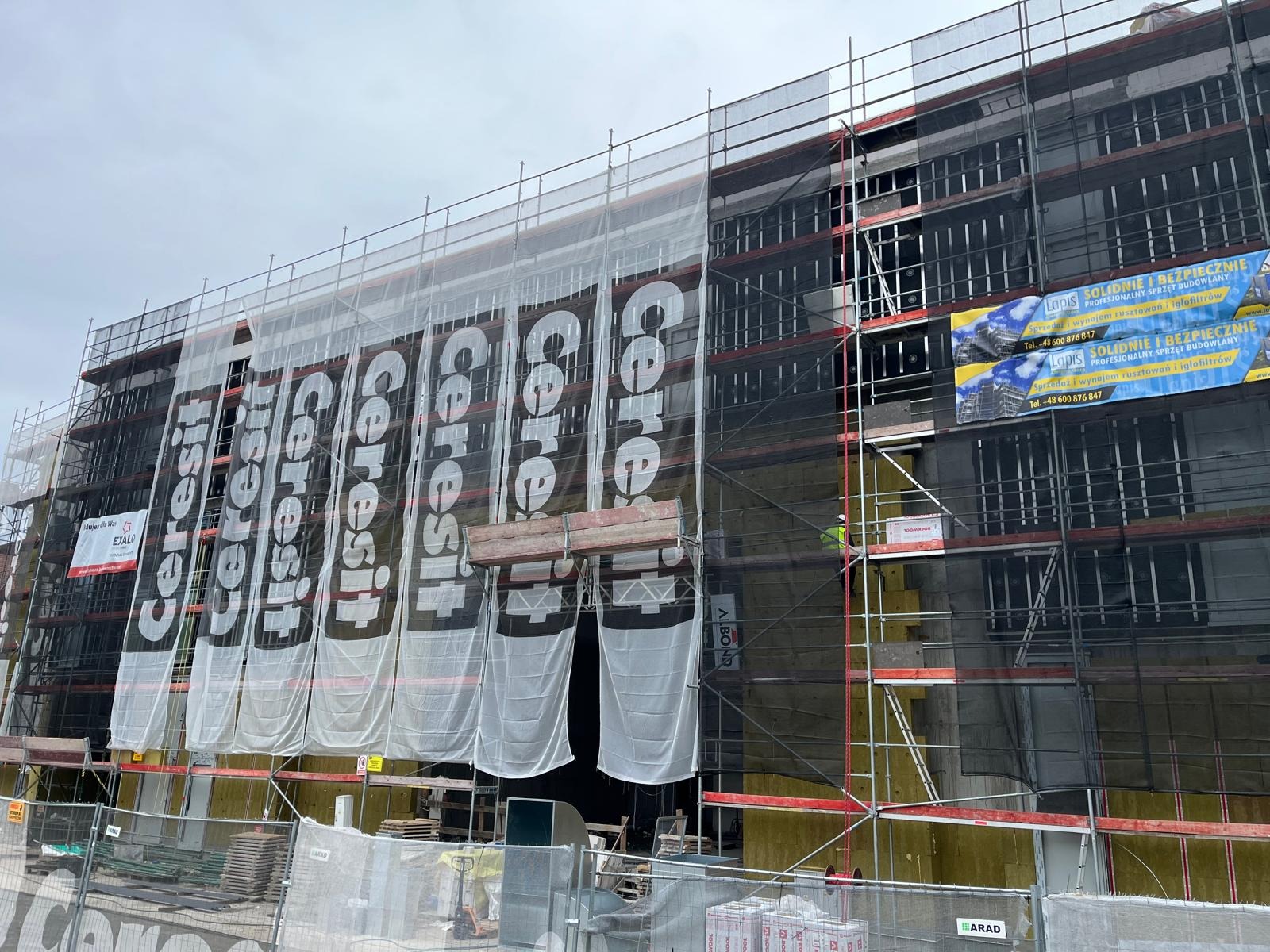 Ocieplanie elewacji budynku wielorodzinnego z użyciem rusztowań, osłonięte banerami reklamowymi Cerestit, widoczne płyty izolacyjne i siatka pod tynk.