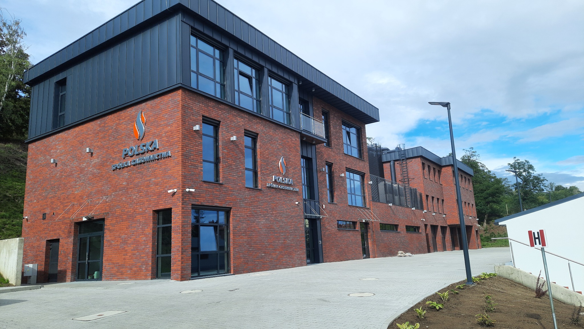 Nowoczesny budynek biurowy z czerwonej cegły z czarnym dachem i dużymi oknami, logo firmy widoczne na elewacji, zadbany chodnik z kostki brukowej.