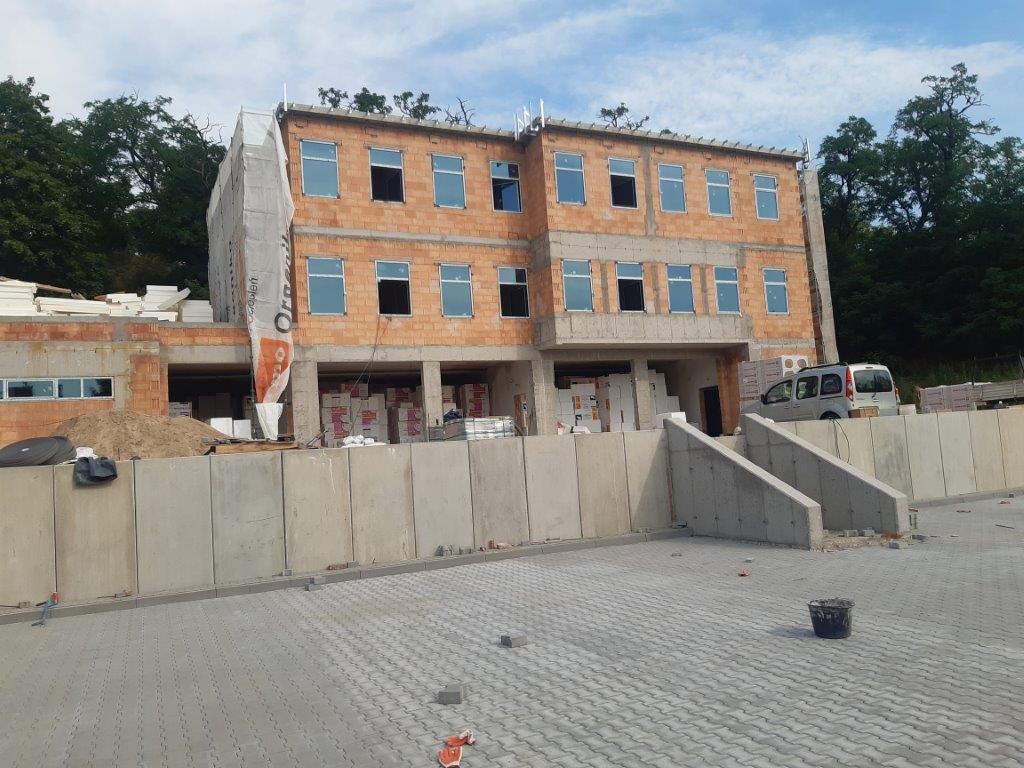 Trzypiętrowy budynek w stanie surowym otwartym, z widocznymi pustakami ceramicznymi i zamontowanymi oknami, częściowo osłonięty plandeką z nadrukiem, z przygotowanym terenem wokół i zaparkowanym...