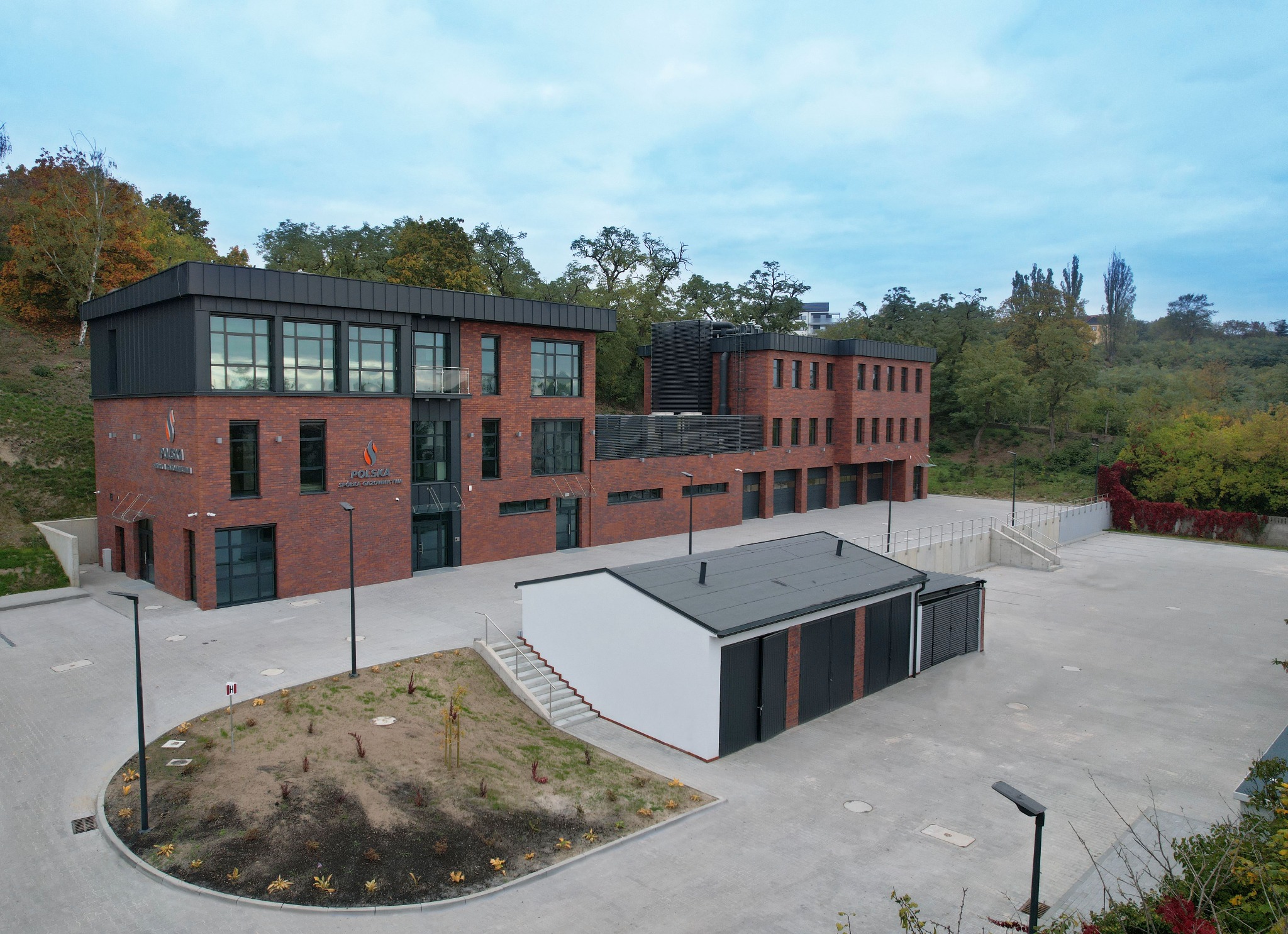 Kompleks budynków biurowych z czerwonej cegły z elementami nowoczesnej architektury, widok z lotu ptaka na parking i otoczenie zieleni.