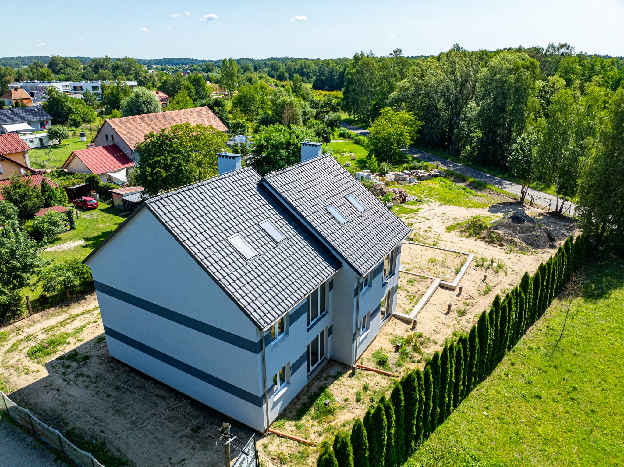 Dwukondygnacyjny dom murowany z szarą elewacją w trakcie budowy, widok z lotu ptaka, z fundamentami i otaczającą zielenią.
