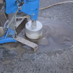 Wiercenie rdzeniowe w asfalcie za pomocą niebieskiej wiertnicy; widoczne chłodziwo i fragment buta pracownika.