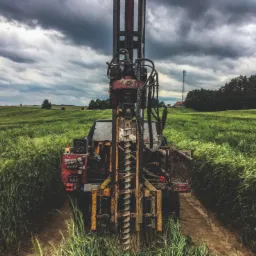 Wiertnica do studni głębinowych na polu, widok z przodu, z widoczną konstrukcją wiertniczą, spiralą wiertła i aparaturą kontrolną, tło: zielone pole i pochmurne niebo.