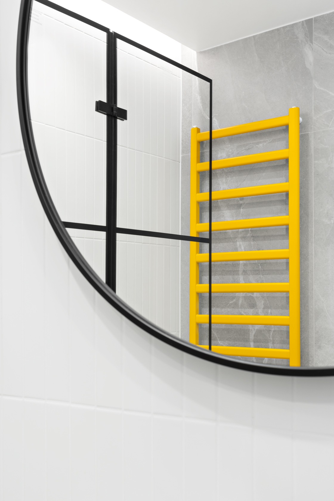 Okrągłe lustro w czarnej ramie odbija żółty grzejnik drabinkowy na tle szarej ściany z efektem marmuru i białą ścianę z płytek w łazience.