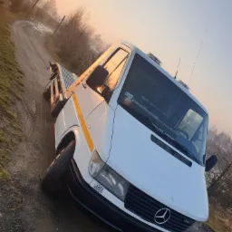 Biała laweta Mercedes-Benz 312D na polnej drodze o wschodzie słońca, z widocznym żółtym pasem na kabinie i charakterystycznym logo Mercedesa na masce.