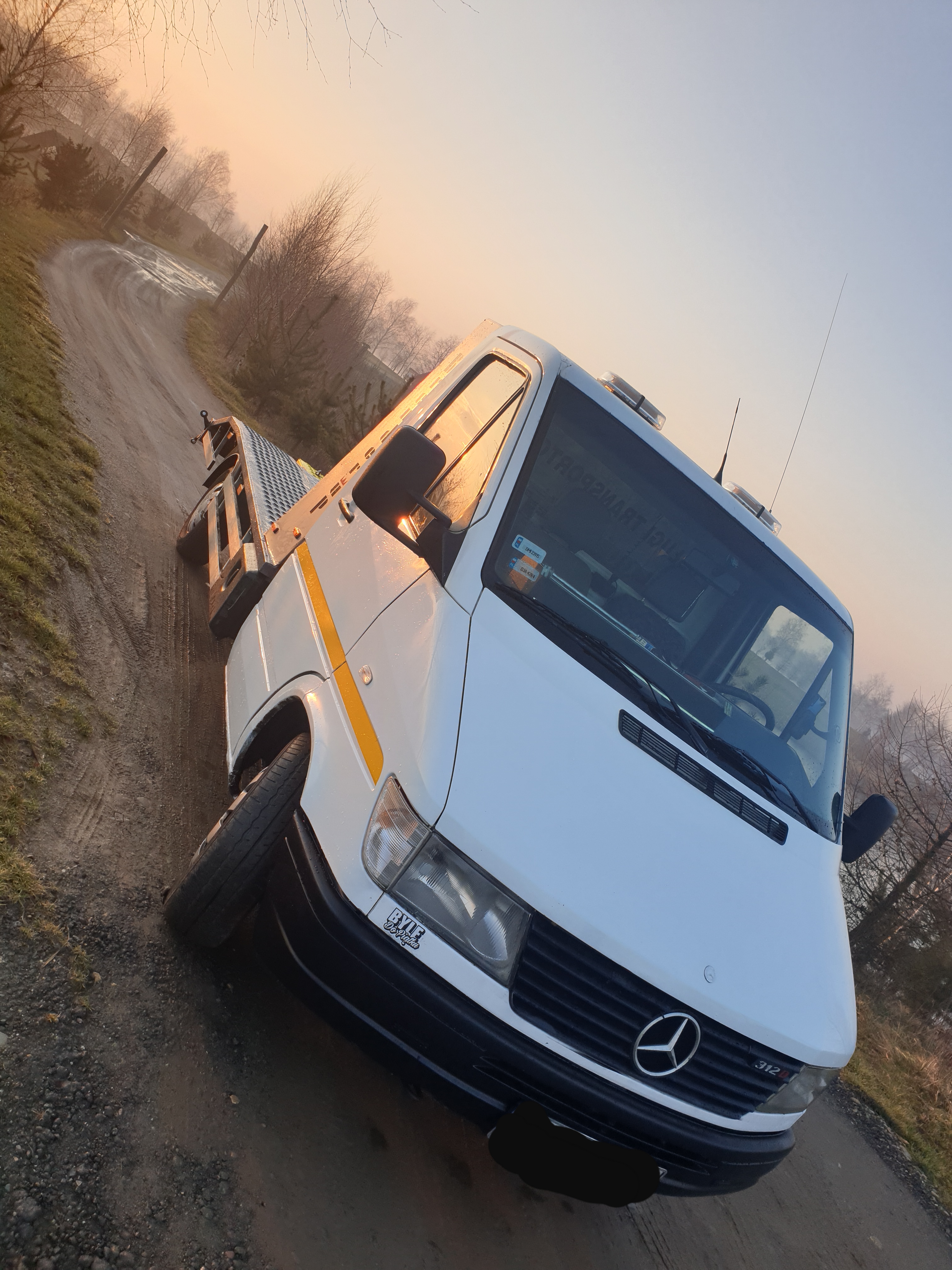 Biała laweta Mercedes-Benz 312D na polnej drodze o wschodzie słońca, z widocznym żółtym pasem na kabinie i charakterystycznym logo Mercedesa na masce.