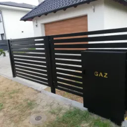 Nowoczesna, czarna brama wjazdowa z poziomymi przęsłami, z widoczną skrzynką gazową z napisem 'GAZ', na tle białego domu z brązową bramą garażową.