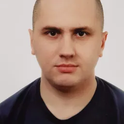 Portret mężczyzny o krótkich włosach w ciemnej koszulce z logo Adidas na jasnym tle. Zdjęcie paszportowe z neutralnym wyrazem twarzy.