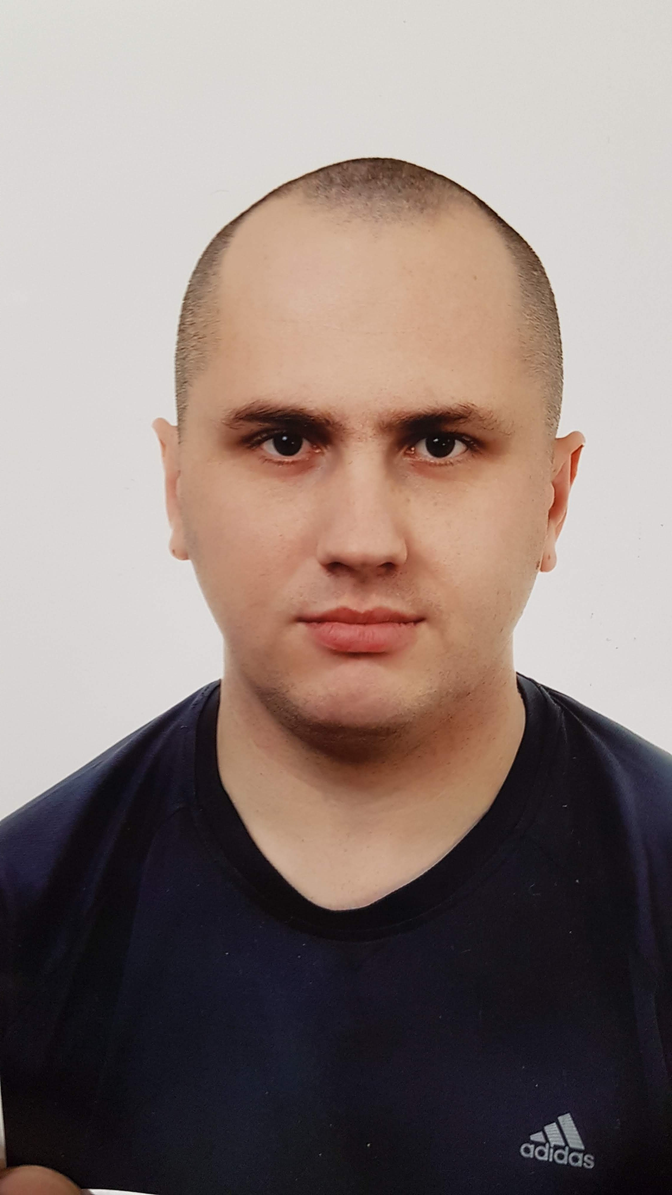 Portret mężczyzny o krótkich włosach w ciemnej koszulce z logo Adidas na jasnym tle. Zdjęcie paszportowe z neutralnym wyrazem twarzy.