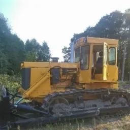 Żółty, gąsienicowy spychacz z otwartymi drzwiami kabiny, stojący na zewnątrz w otoczeniu drzew.