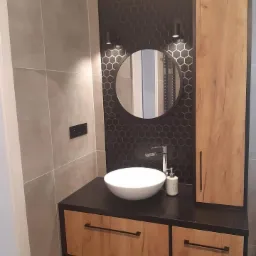 Nowoczesna łazienka z okrągłym lustrem podświetlonym kinkietami, umywalką nablatową i szafką z drewnopodobnym frontem, szare płytki i heksagonalna tapeta.