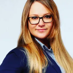 Portret uśmiechniętej blondynki w okularach i granatowej marynarce na jasnym tle.