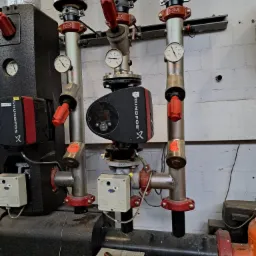 Węzeł cieplny z pompami obiegowymi Grundfos, rury stalowe z izolacją termiczną, manometry i zawory z czerwonymi pokrętłami na tle ściany z białej cegły, klucz nasadowy leżący na drabinie.