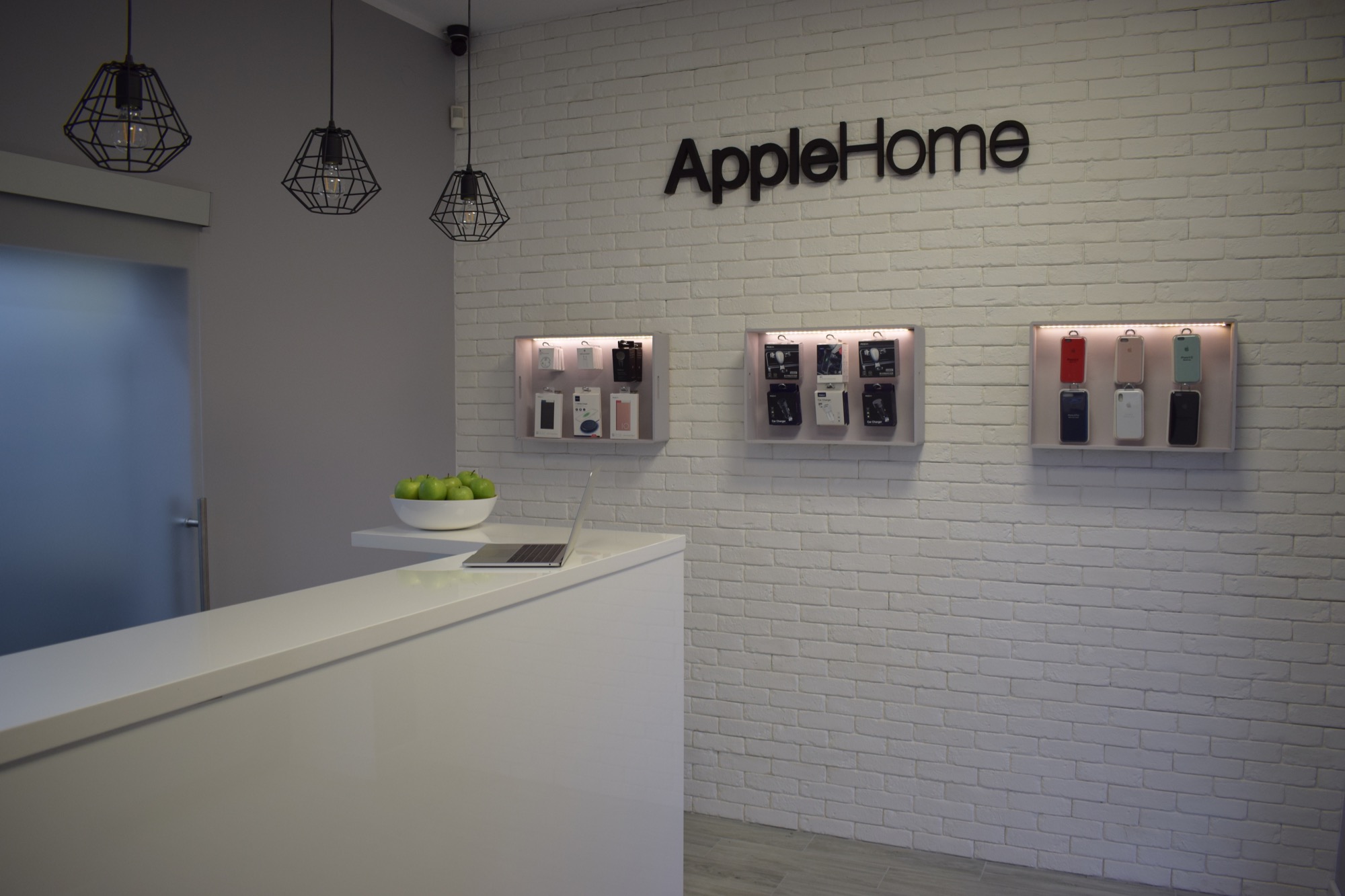 Wnętrze sklepu AppleHome w Warszawie z ladą, laptopem, misą zielonych jabłek i ekspozycją akcesoriów na białej ceglanej ścianie pod trzema lampami w stylu industrialnym.