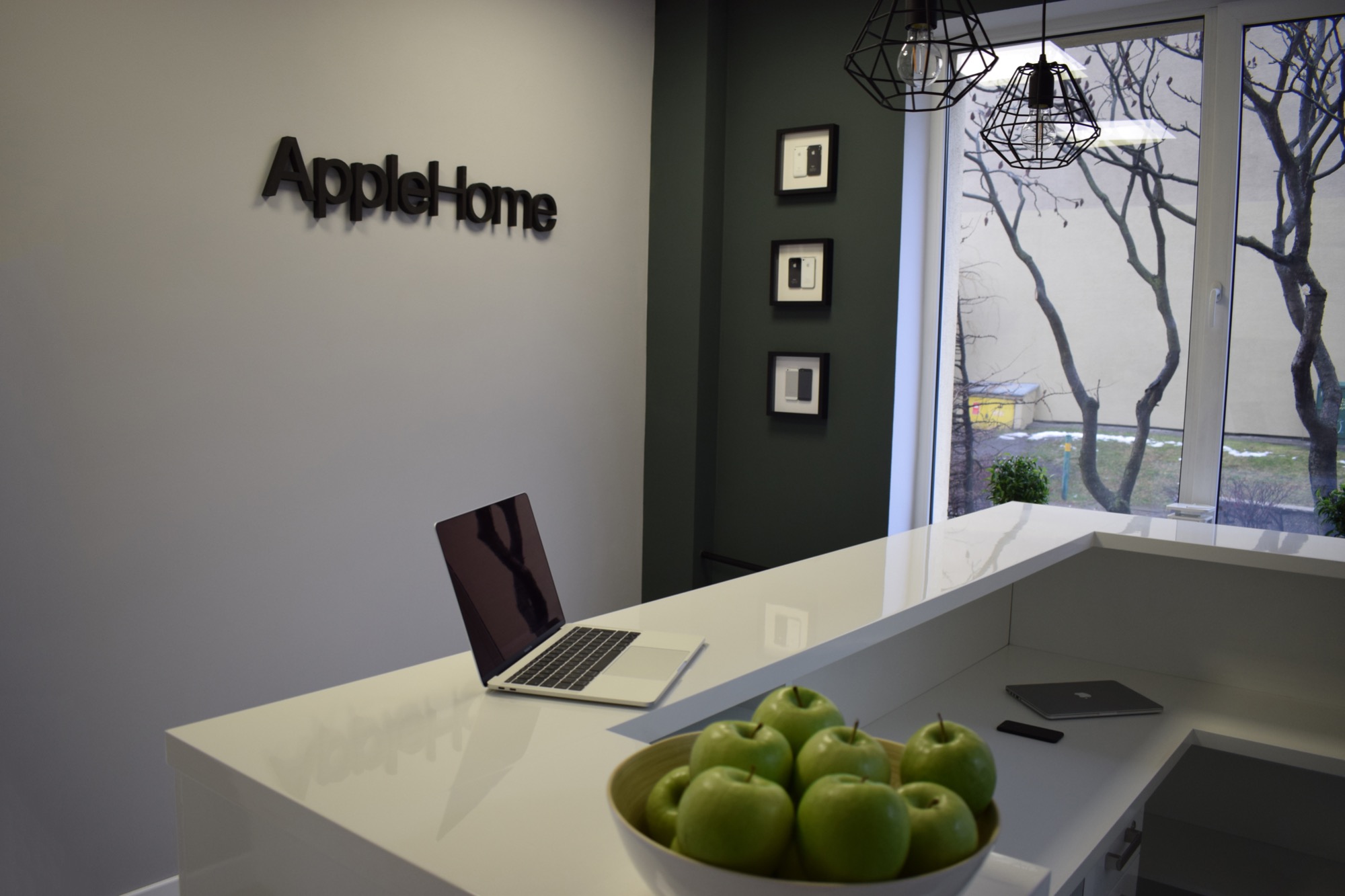 Recepcja firmy z laptopem i misą zielonych jabłek na blacie, na ścianie logo firmy AppleHome i zdjęcia telefonów w ramkach